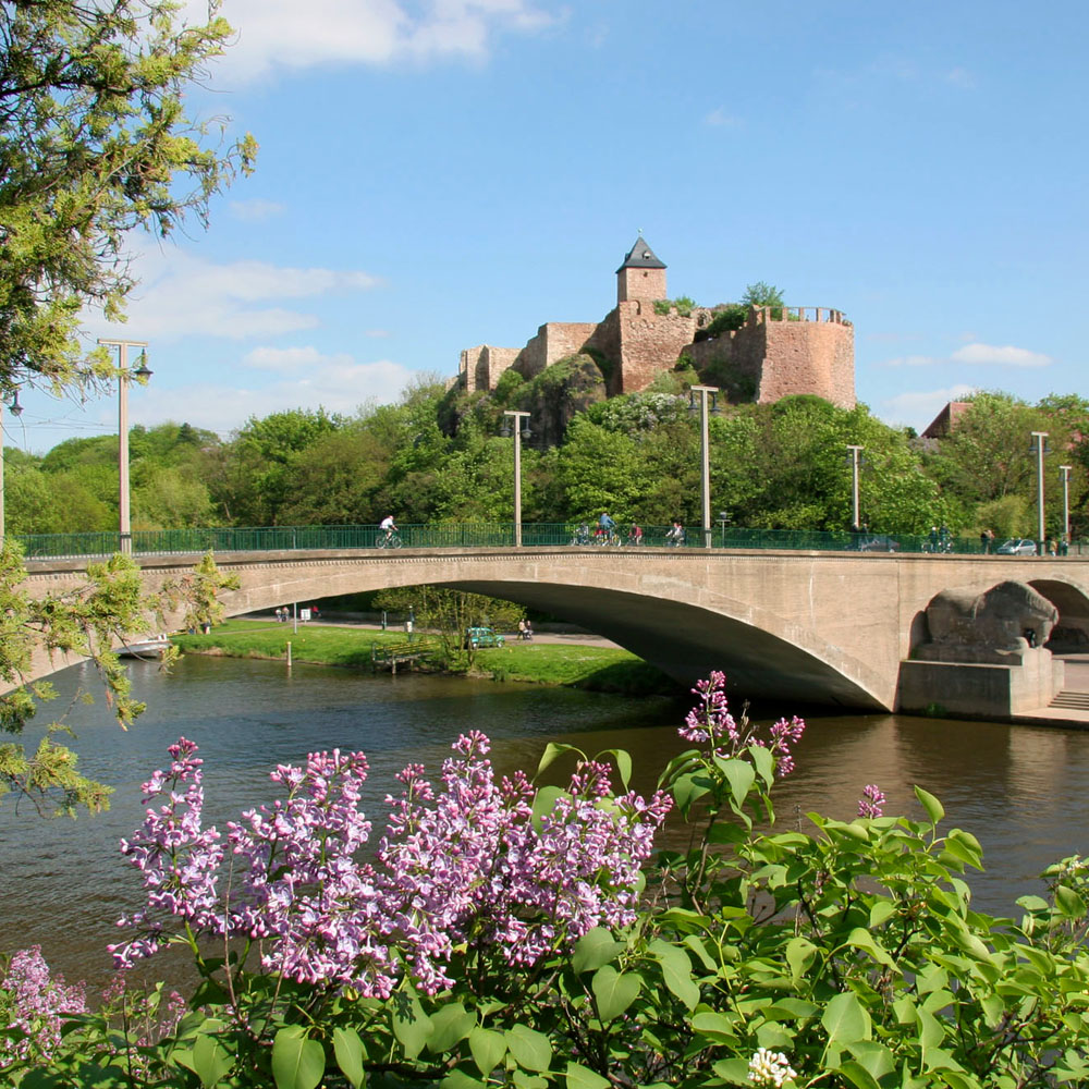 Ansicht einer sommerlichen Landschaft mit Steinbrücke auf der in der Entfernung die Burg Giebichenstein zu sehen ist.